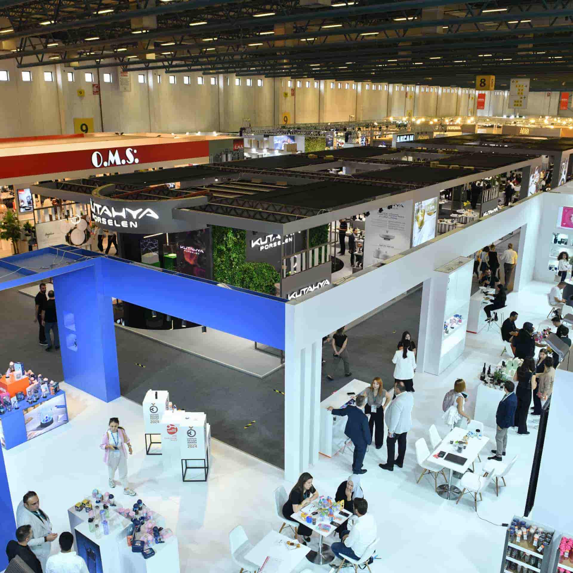 Katılımcı stantları, ürünleri keşfeden ve sohbet eden ziyaretçilerle dolu salonun kuş bakışı görünümü.
